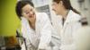 Two young female researchers in white lab coats and protective gloves looking at each other and smiling as they work on a project.